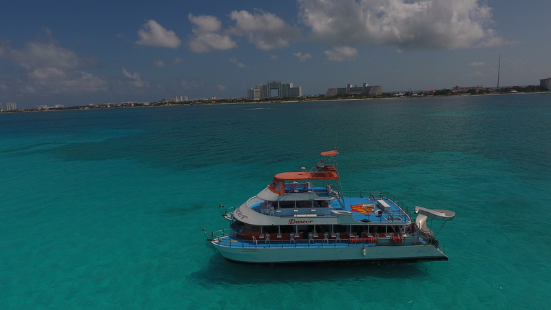 Vista aérea del catamarán de fiesta Dancer de tres pisos anclado sobre agua turquesa con la silueta de la zona hotelera de Cancún al fondo