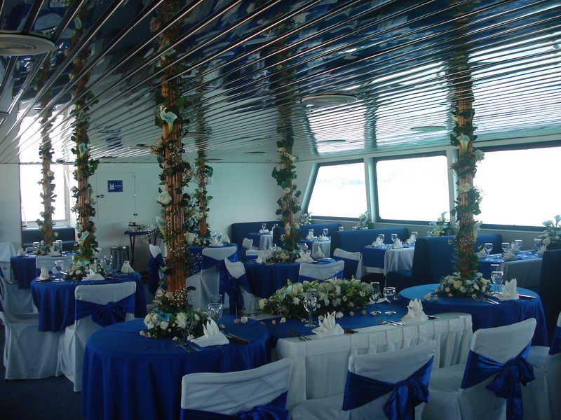 Interior salon decorated for a wedding: round tables with royal-blue cloths and white chair covers with blue sashes, support columns wrapped in ivy and white calla lilies, mirrored ceiling reflecting the setup.