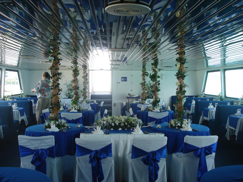 Symmetric interior view of the salon set for a wedding, blue and white round tables, columns wrapped in greenery and calla lilies, a low white floral centerpiece on the head table, large windows and mirrored ceiling.
