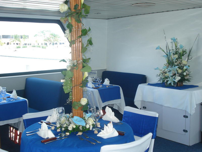 Set dining table aboard the boat with royal-blue cloth, white napkins and a column wrapped in greenery and roses beside it; a blue floral arrangement on a side cabinet, window showing palms and marina outside.