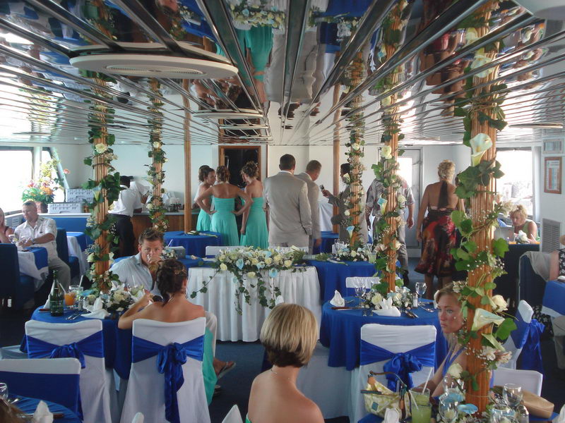 Bride in a white gown and groom in a tan suit posing together standing on the white bow of the boat, brilliant turquoise sea and blue sky behind, rigging lines to the side.