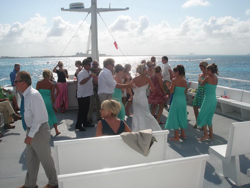 Guests dancing on the open upper deck around the Furuno mast, women in teal dresses, sunlit shimmering sea and Cancun skyline faint on the horizon, white benches around the deck edge.