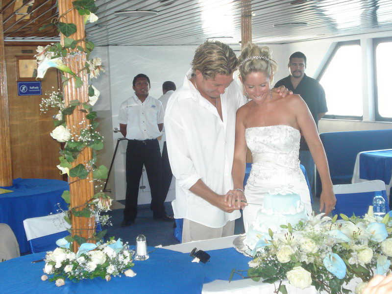 Bride and groom cutting a pale-blue tiered wedding cake indoors, two crew members watching in the background, ivy-wrapped column and blue floral-decorated tables around them.