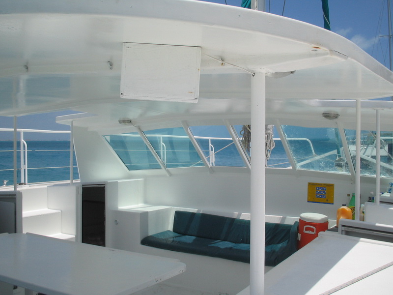 Shaded seating area on the deck of a white sailing catamaran, cushioned bench, tables, a red drink cooler and juice bottles, bright open sea visible through the open sides on a sunny day.