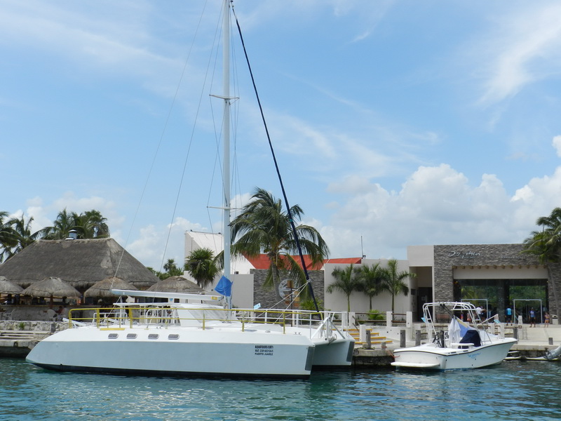 White sailing catamaran ('Aquatours Cat') moored at a marina beside a small center-console panga, palm trees and a stone dive-shop building behind, sunny with puffy clouds.