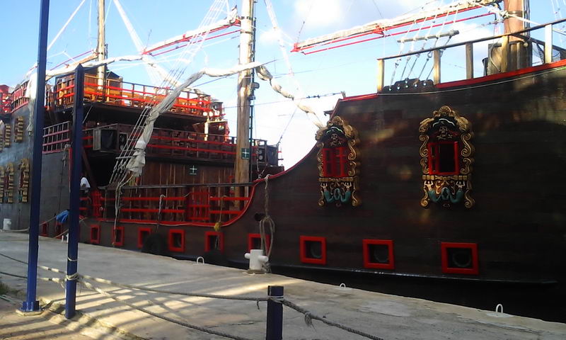 Casco lateral del galeón Jolly Roger en el muelle con marcos dorados en las troneras, ribetes rojos y velas recogidas en sus altos mástiles