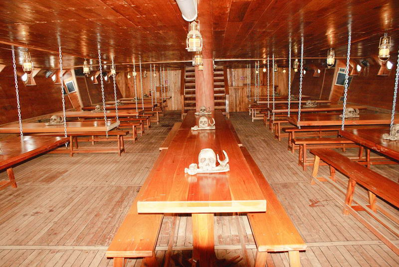 Comedor bajo cubierta del galeón Jolly Roger con largas mesas barnizadas, bancas, lámparas colgantes y calaveras decorativas