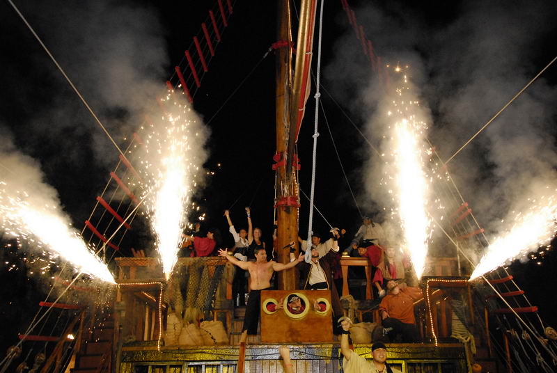 Final nocturno del show pirata en el galeón Jolly Roger con fuentes de fuegos artificiales brotando del aparejo mientras la tripulación celebra