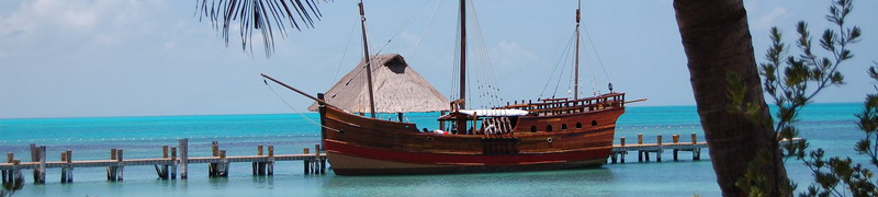 Réplica de carabela española amarrada junto a un muelle de madera en el agua turquesa del Caribe cerca de Isla Contoy, enmarcada por hojas de palmera