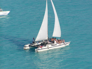 Aerial photo of a white sailing catamaran with two raised white sails and several people aboard, crossing clear turquoise water with the bow of another boat visible at the upper left.