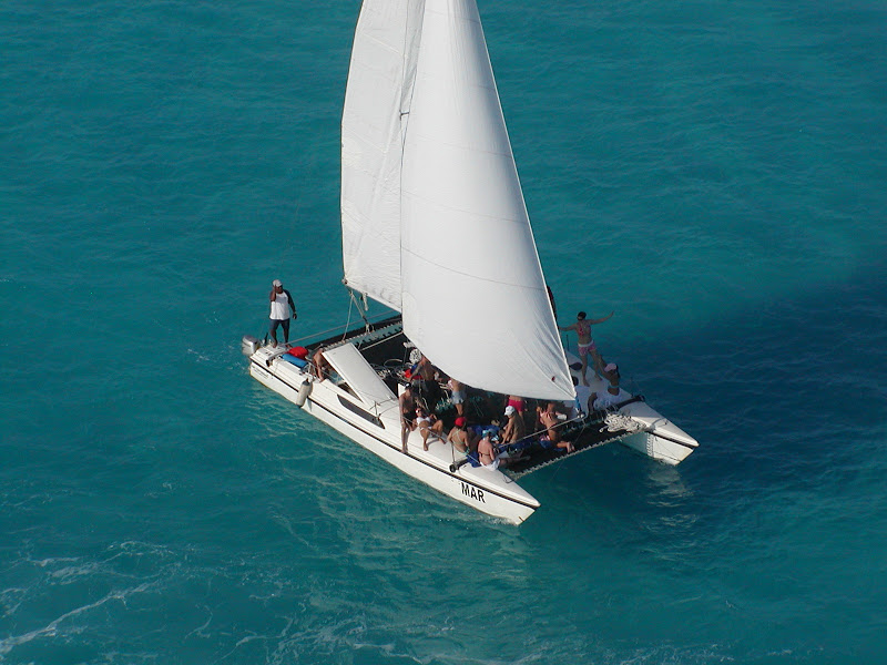 Aerial view of a white sailing catamaran marked 'MAR' under mainsail, carrying a group of passengers on deck, crossing open turquoise sea on a sunny day.
