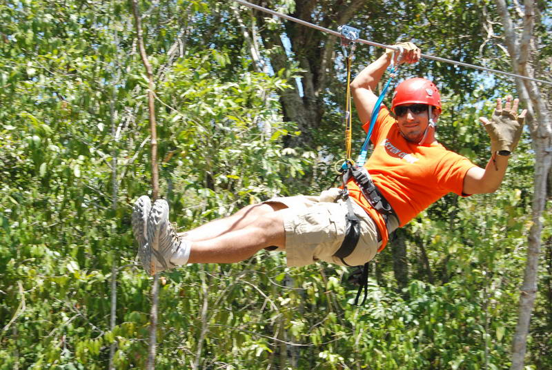 Visitante con casco y camisa naranja deslizándose en una tirolesa de dosel sobre la selva verde en el parque Aktun Chen, 