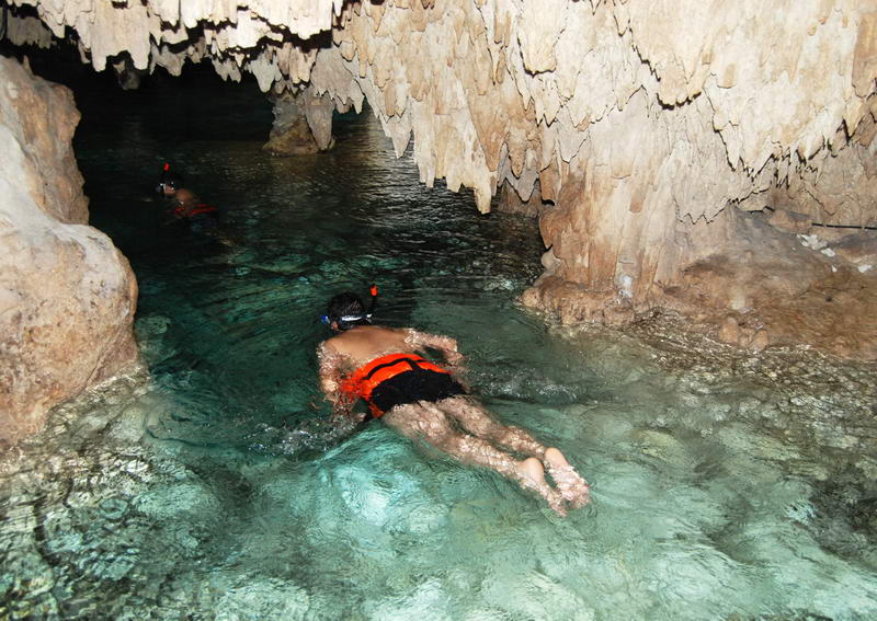 Persona con chaleco salvavidas naranja nadando por un estrecho pasaje de estalactitas en el cenote subterráneo de Aktun Chen
