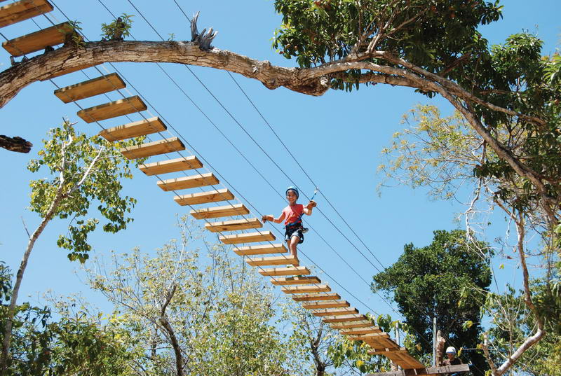 Niño cruzando un puente colgante de madera entre los árboles de la selva en el circuito de dosel de Aktun Chen bajo un cielo azul