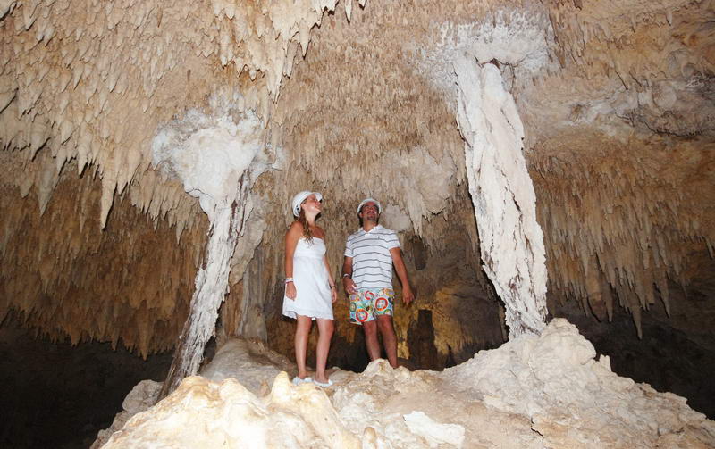Pareja de pie sobre una formación rocosa dentro de la enorme cueva seca de Aktun Chen, contemplando el techo cubierto de estalactitas
