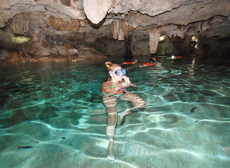 Persona haciendo esnórquel en el agua turquesa cristalina del cenote subterráneo de la cueva Aktun Chen, con estalactitas sobre su cabeza