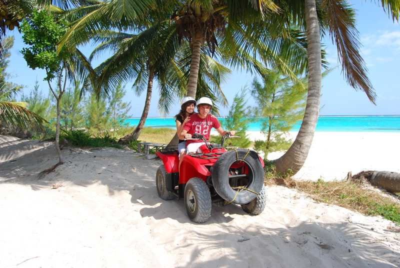 Pareja con cascos en una cuatrimoto roja estacionada bajo las palmeras junto a la arena blanca y el agua turquesa de Playa Maroma