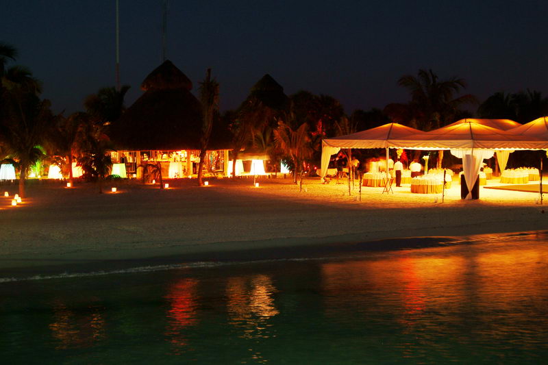 Nighttime beach event scene with a thatched palapa bar glowing warm orange, a white catering tent, and candle luminaries lining the sand, their light reflecting on the calm shallow water.