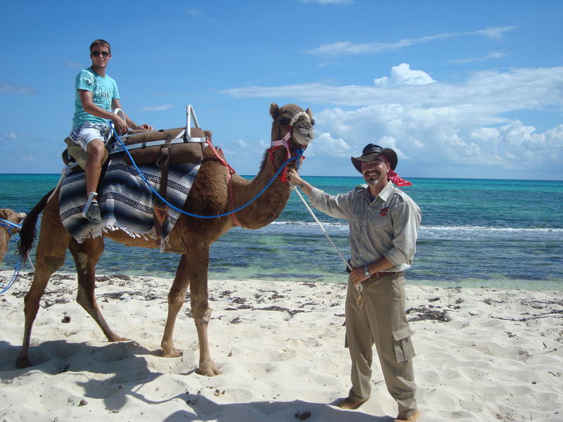 Joven montando un camello guiado por un guía sonriente por la playa de arena blanca junto al Caribe turquesa en Maroma