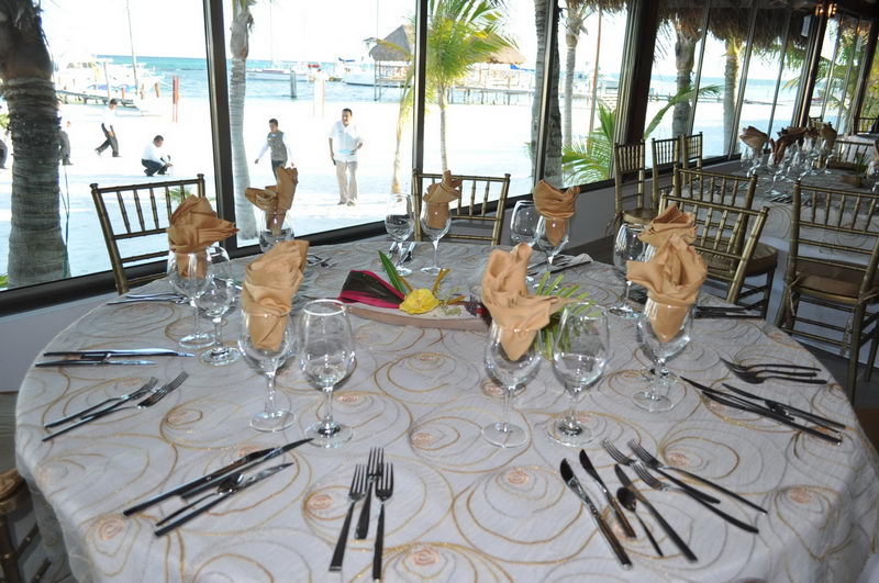 Mesa elegante con servilletas doradas y vista al muelle y las embarcaciones de Maroma desde el restaurante frente al mar, Riviera Maya