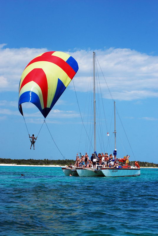 A person swings on a rope beneath a colorful red, yellow and blue parachute/spinnaker hanging off a white sailing catamaran packed with cheering passengers. Bright turquoise sea, a sandy islet, and clear blue sky behind.