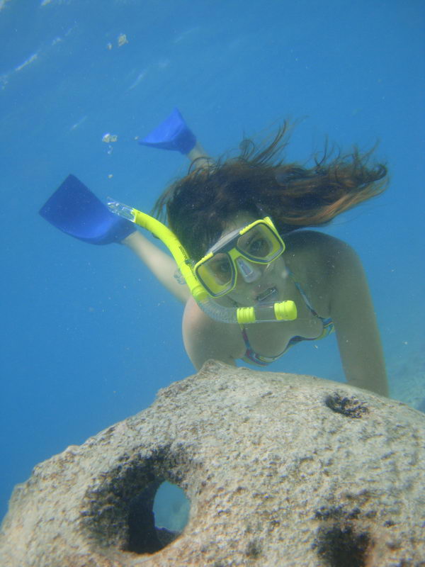 Underwater shot of a young woman snorkeling in clear blue water, wearing a yellow mask and snorkel and blue fins, hair floating upward. A rock with holes sits in the foreground below her.