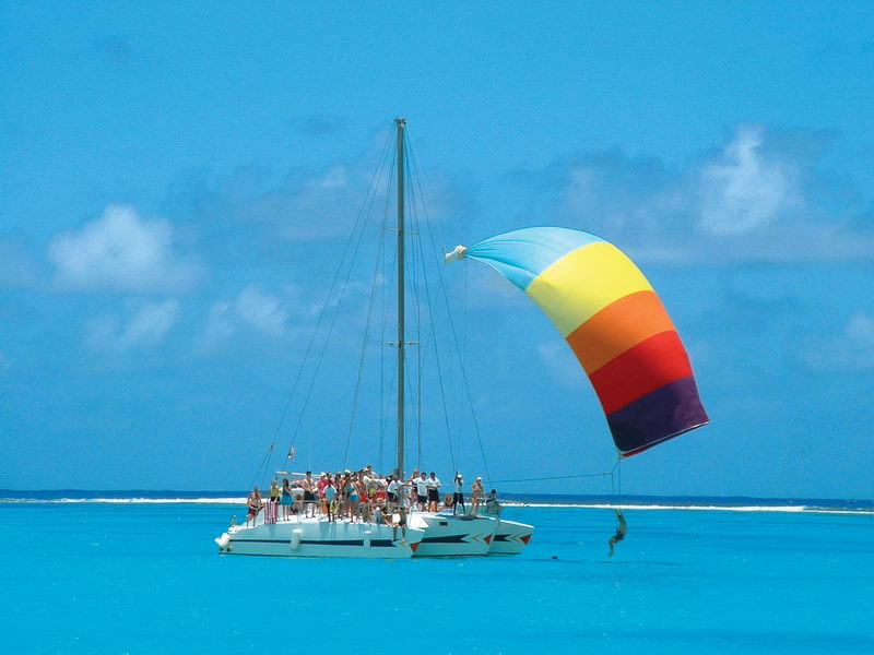 A white sailing catamaran full of passengers floats on brilliant turquoise water, deploying a large rainbow-striped spinnaker sail (blue, yellow, orange, red, purple) against a blue sky with scattered clouds.