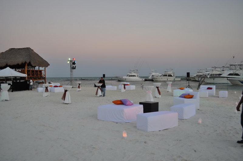 Montaje de evento al atardecer en la playa de Maroma con salas lounge blancas, cojines de colores y yates al fondo, Riviera Maya