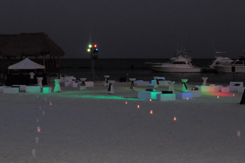 Montaje nocturno de evento en la playa con iluminación LED de colores, velas y yates de pesca deportiva atracados en Marina Maroma, Riviera Maya