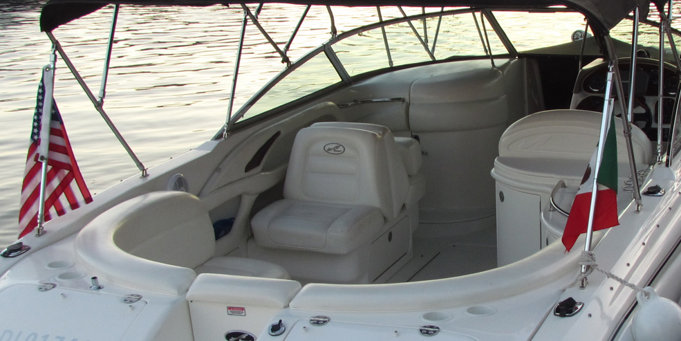 Cockpit of a white Sea Ray sport cruiser with tan/cream upholstered helm seat and lounge, bimini frame overhead, US and Mexican flags on the stern rails. Calm water at sunset behind. Empty seats, tidy layout.