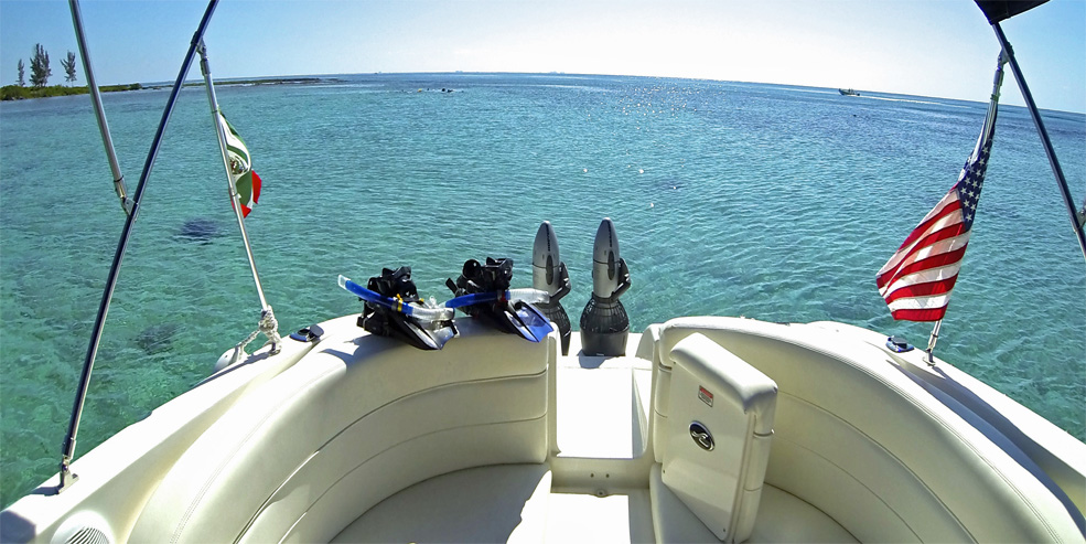 Wide-angle view from the bow of a Sea Ray cruiser looking out over calm clear turquoise water toward a reef and horizon. Two sets of snorkel fins and gear rest on the cream bow seating; US and Mexican flags on the rails. Sunny.