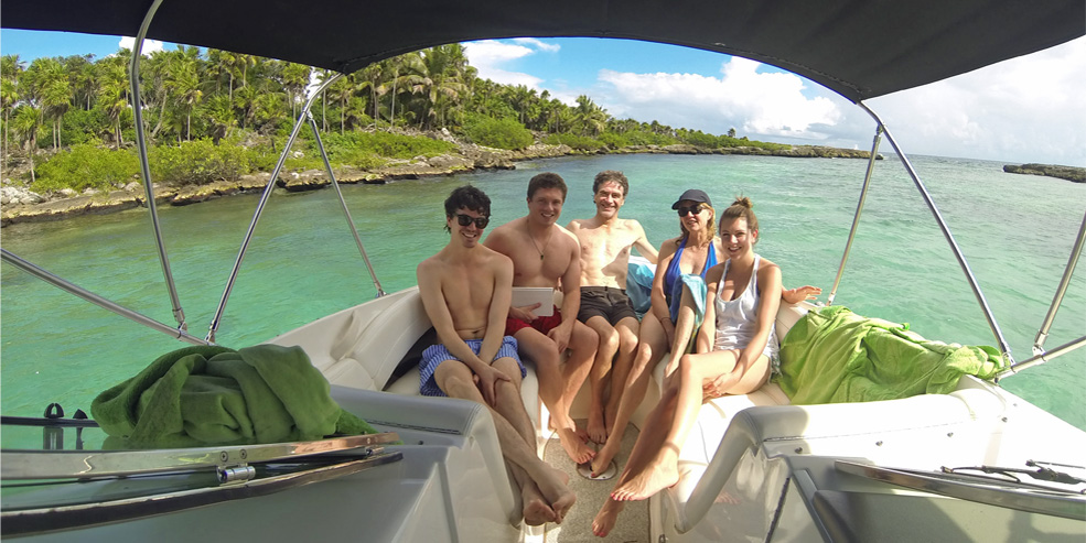 Five smiling young people in swimwear sit together on the cushioned bow/cockpit of a small cruiser under a bimini top, anchored in calm turquoise water beside a rocky jungle-lined inlet. Green towels, sunny day, relaxed group.