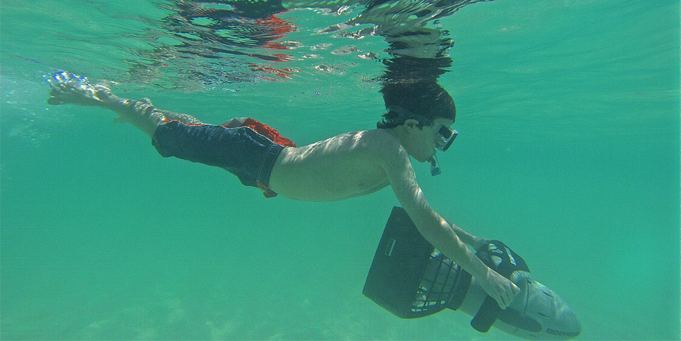 Underwater shot of a boy snorkeling near the surface in green-tinted clear water while holding onto a black electric underwater sea scooter (diver propulsion vehicle). Sandy bottom below, another swimmer above. Sunny shallows.