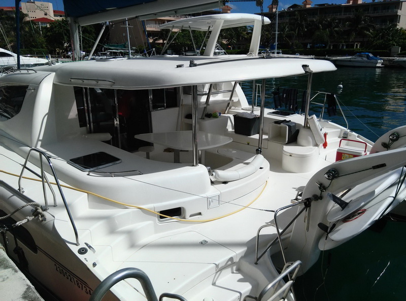 Aft view of a white Leopard sailing catamaran docked stern-to, showing the hard bimini top over the cockpit, dinette table and seating, twin transoms with a tender/dinghy on the port side. Clear water, palms and resort behind, sunny.