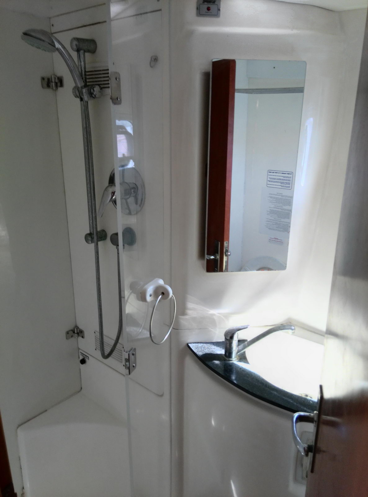 Compact boat head/bathroom with white fiberglass walls, a handheld shower on a slide rail, a small corner sink with single faucet, a mirrored wood cabinet and a towel ring. Cramped marine wet-head layout.