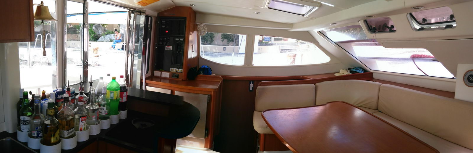 Wide interior view of a catamaran salon and galley, cream leather seating around a mahogany table, electrical breaker panel on the bulkhead, open patio doors to a dock, and a bar area lined with liquor bottles.