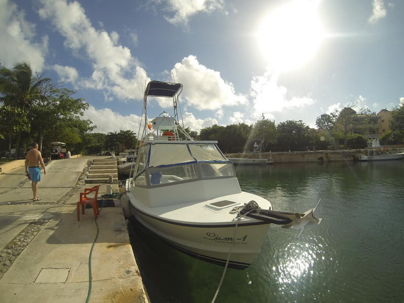 Fisheye view of a white sportfishing boat named Liam-1 moored bow-out along a concrete dock at sunny golden hour, a shirtless man walking on the quay, canal and buildings in the background.