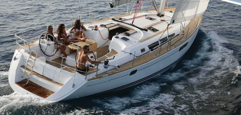 Stern-quarter view of a Jeanneau Sun Odyssey 49 under sail with four people aboard: man at one of the twin wheels, three guests seated around the teak cockpit table, fold-down swim platform, gentle sea.
