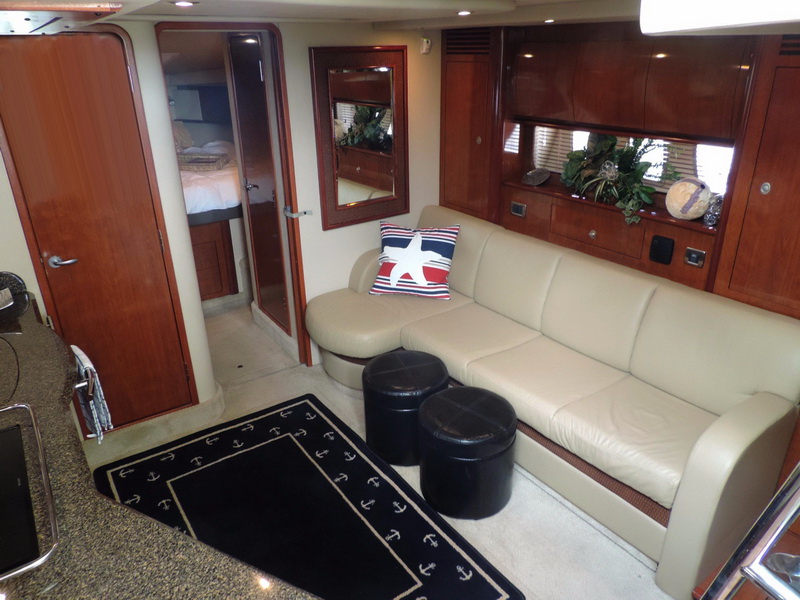 Sea Ray yacht salon with tan leather L-shaped sofa, nautical star pillow, black ottomans, anchor-pattern rug, cherry wood cabinetry, mirror and doorway to forward cabin with bed visible.