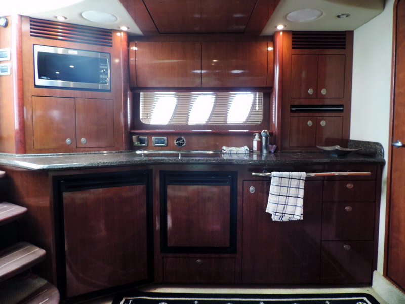 Yacht galley with dark cherry cabinetry, granite countertop, stainless microwave, sink, refrigerator and dish towel on rail, three windows with blinds above the counter.