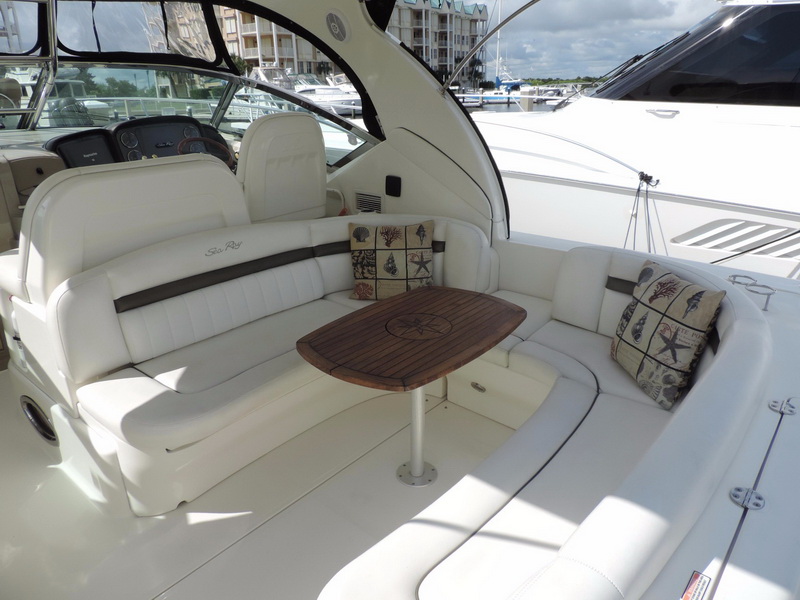 Sea Ray cockpit with wraparound white leather seating, wooden pedestal table with compass-rose inlay, decorative seashell pillows, helm station at left and marina condos in the background.