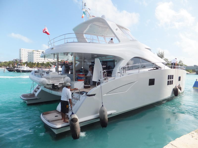 Large white power catamaran docked stern-to at a stone quay, crew member handling lines beside black fenders, flybridge with flags above, hotel and tour boats in background, bright turquoise water.