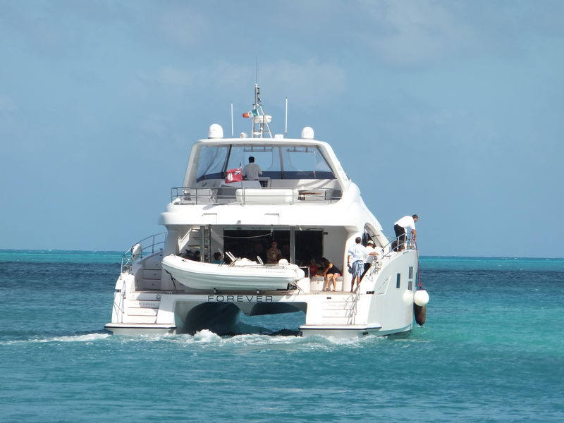 Stern view of power catamaran FOREVER motoring away with guests in the open aft salon, crew standing on the side deck, gray tender on davits between the hulls, reef line and turquoise bands of sea behind.