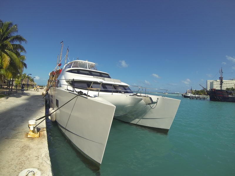 Bow view of a large white power catamaran moored along a palm-lined stone quay, wide twin hulls with mooring lines, pirate-style galleon ship and hotel visible across the channel, deep blue sky.