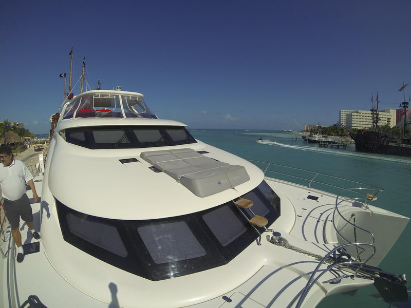 Looking aft along the coach roof of a power catamaran with a gray sunpad and curved white superstructure, man in white polo walking the side deck, turquoise lagoon, dock and pirate ship in the distance.
