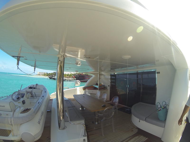 Covered aft deck of a power catamaran with long teak dining table, transparent ghost chairs, gray cushioned lounger with turquoise bag, and a white Williams jet tender parked on the platform, sea views both sides.