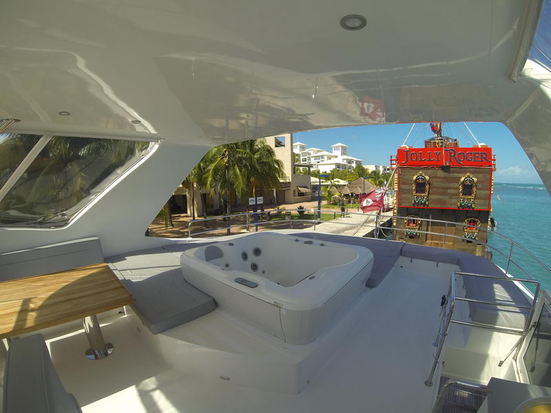 Flybridge of a catamaran with white jacuzzi tub surrounded by gray sunpads and teak table, overlooking a dock with the red Jolly Roger pirate ship attraction, palm trees and condos behind, sunny day.