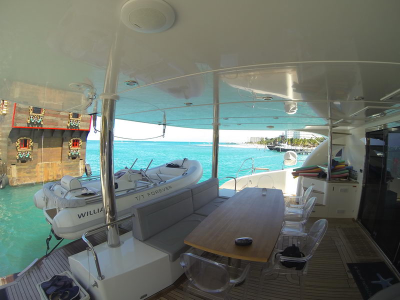 Aft deck of catamaran FOREVER with teak dining table and clear acrylic chairs, gray sofa, rolled colorful pool noodles on the side unit, Williams tender T/T FOREVER on the platform, turquoise bay and hotels behind.