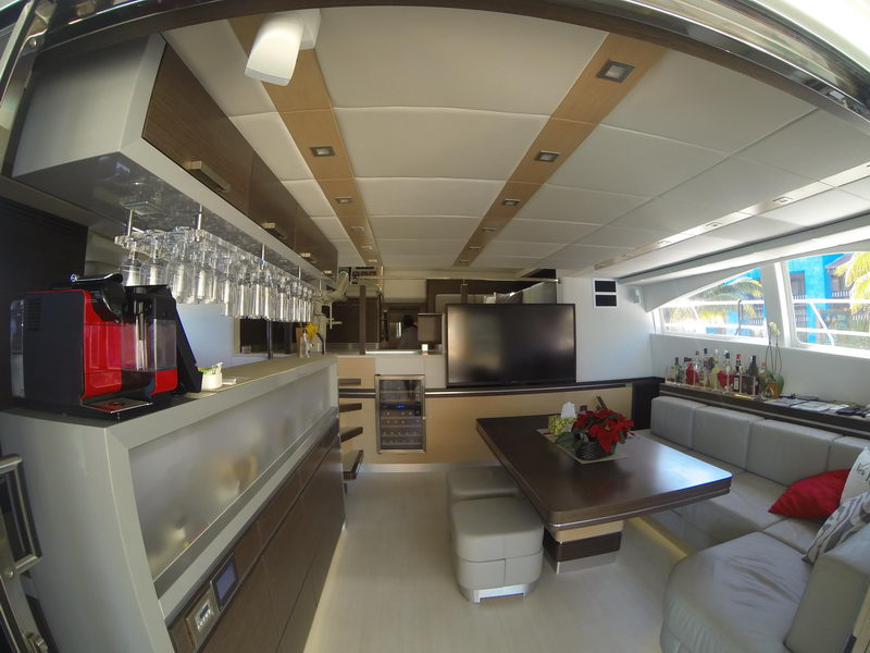 Modern catamaran salon with illuminated bar, rows of hanging stemware, red espresso machine, wine fridge, large flat TV, gray sectional sofa with red cushions, liquor bottles along the window shelf.