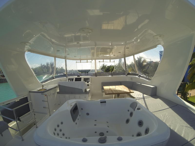 Enclosed flybridge of a power catamaran with white jacuzzi in the foreground, helm station with instruments, wraparound gray upholstered seating and small table, clear roll-down enclosure panels, marina views.
